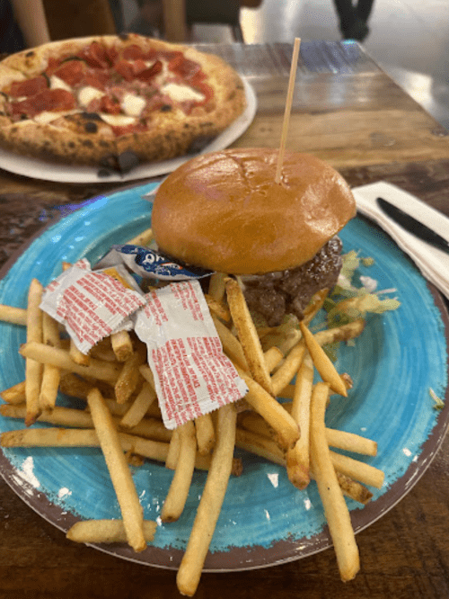 A burger with lettuce and condiments on a blue plate, accompanied by a side of fries and a pizza in the background.